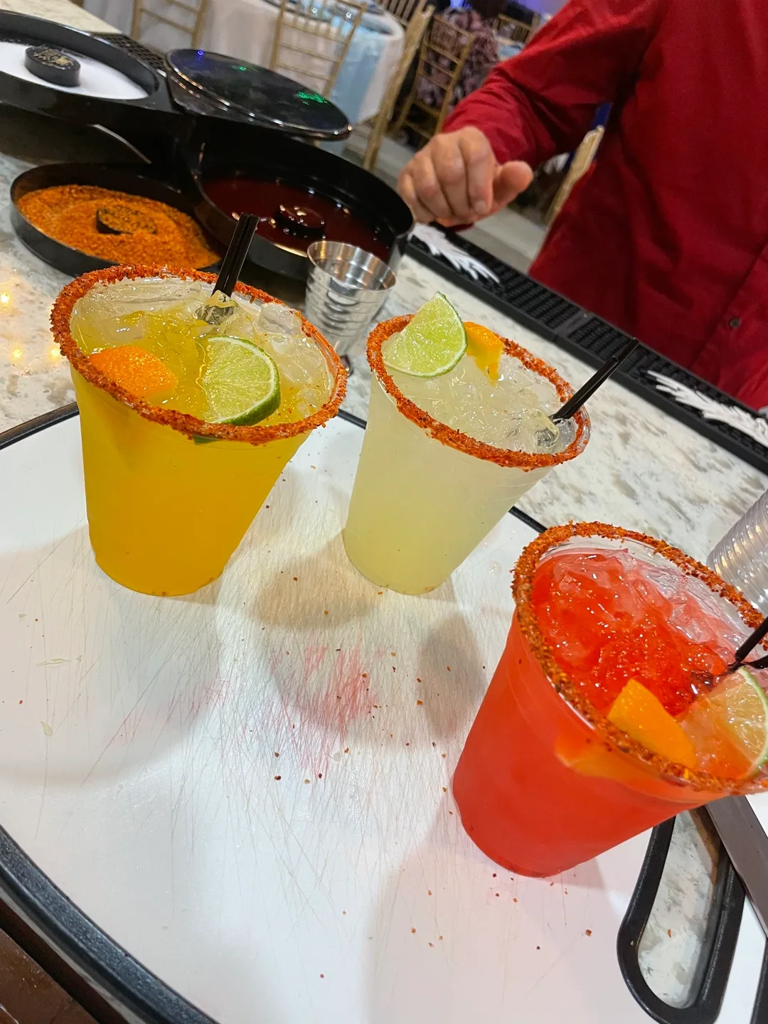 Three chili-rimmed cocktails on a white board — a golden margarita, a pale lime drink and a coral-red one, each packed with ice and garnished with a lime wheel or orange slice, with a bartender in a red shirt at the bar behind