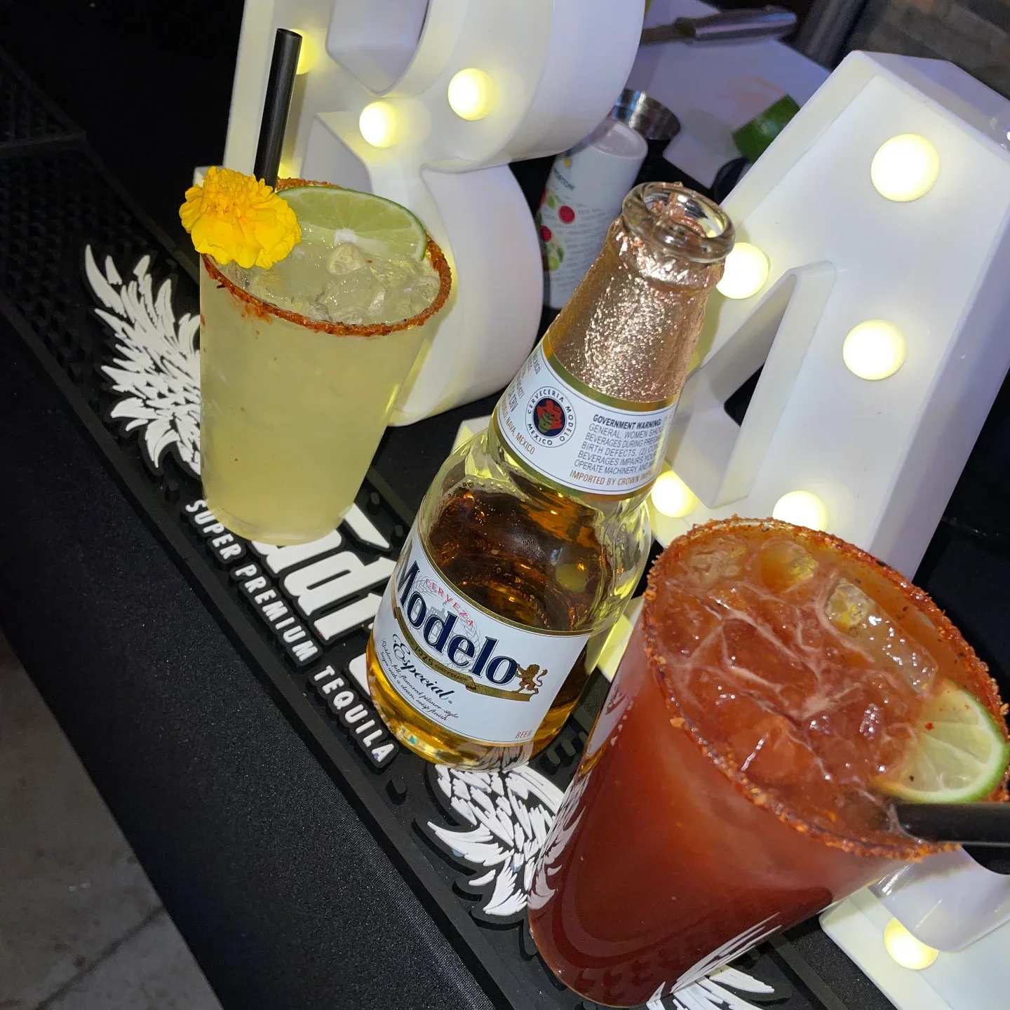 A margarita with a chili rim and yellow flower, an open Modelo bottle and a chili-rimmed michelada in a row on a black tequila mat, glowing marquee numbers behind
