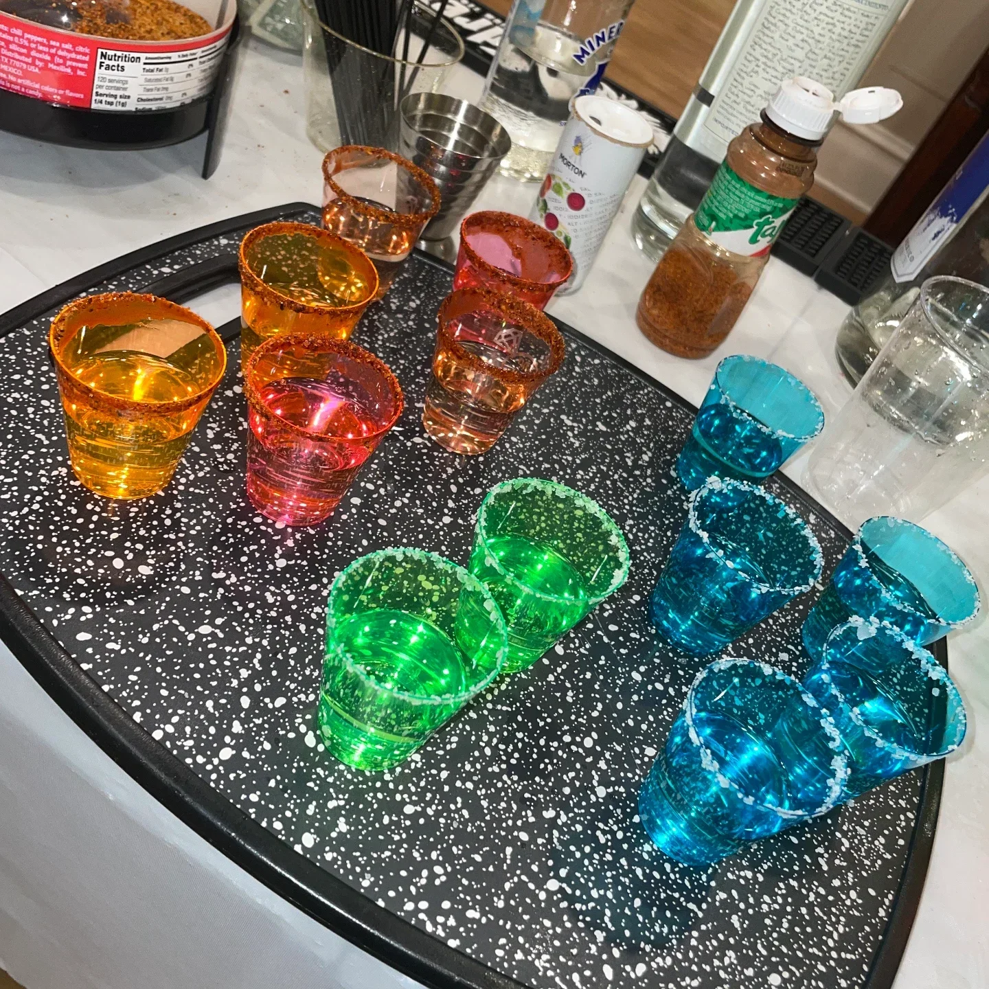 Twelve neon shot glasses in yellow, pink, green and blue on a speckled board, rimmed with chili or salt, with seasoning, salt, club soda and a tequila bottle behind
