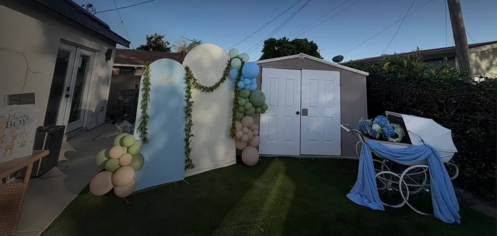 Arch & Balloon Décor: Blue and cream arch backdrops with sage, blue and nude balloon clusters, gold “Liam” lettering and a vintage white carriage on a lawn