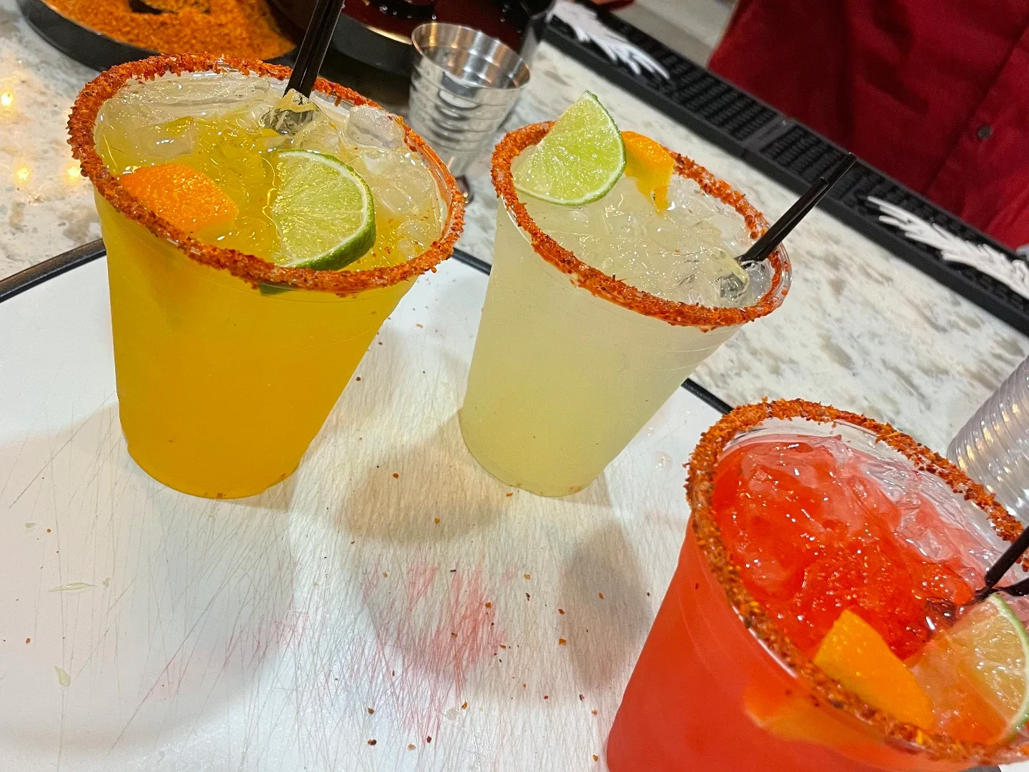Bartender Service: Three chili-rimmed cocktails on a white board — golden, pale lime and coral-red, with lime wheels, orange slices and black straws