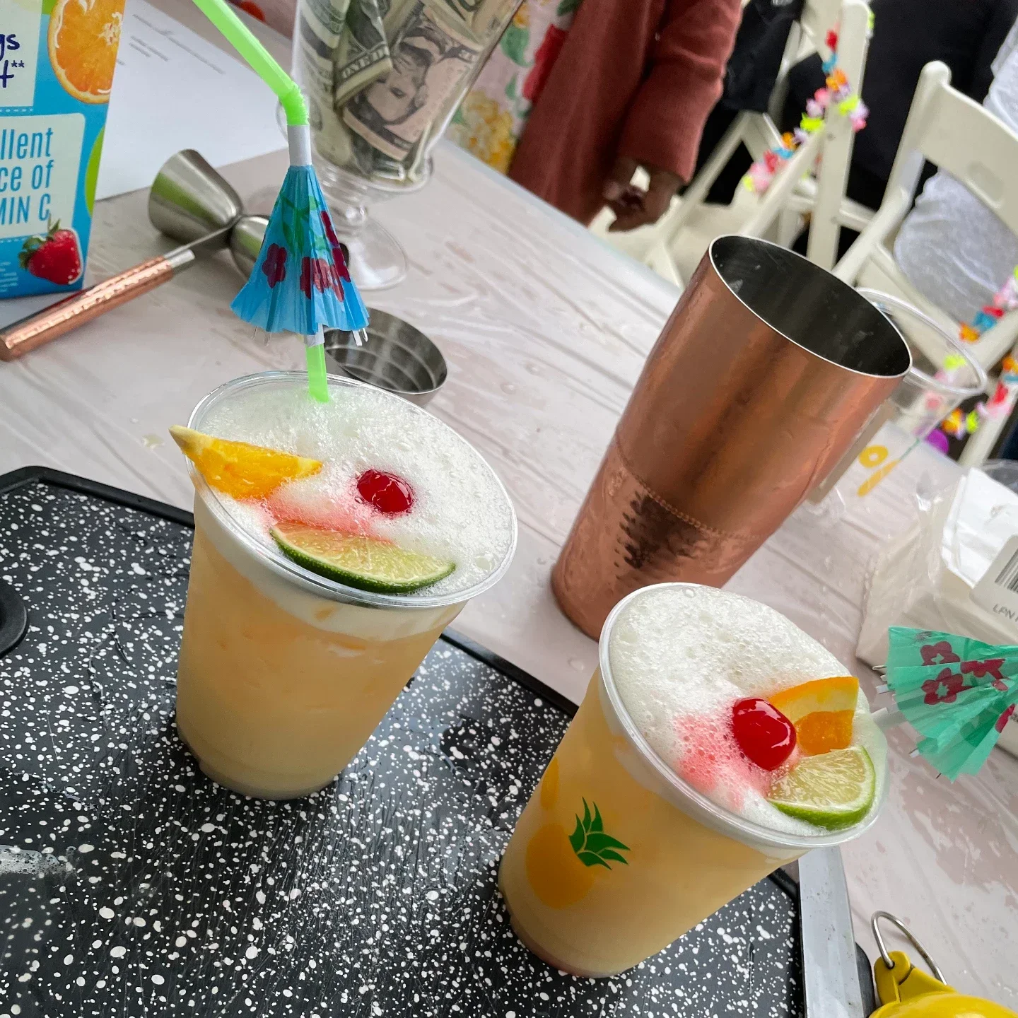 Two foamy tropical cocktails with orange slices, lime wedges, cherries and paper umbrellas, a copper shaker and jigger beside them on a bar table