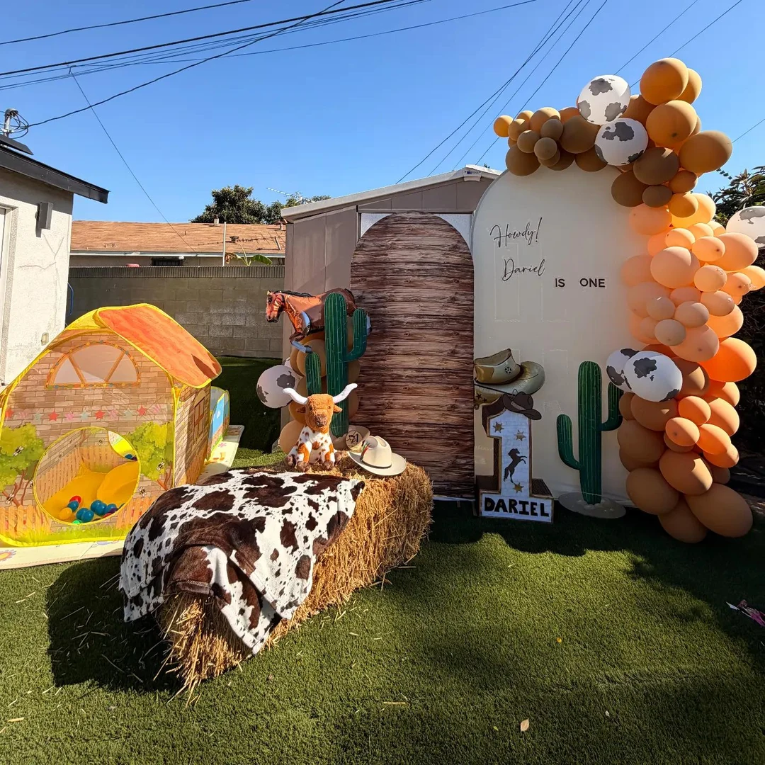 Little cowboy first-birthday backdrop with orange and brown balloons, cow-print accents, cactus cutouts and hay bales