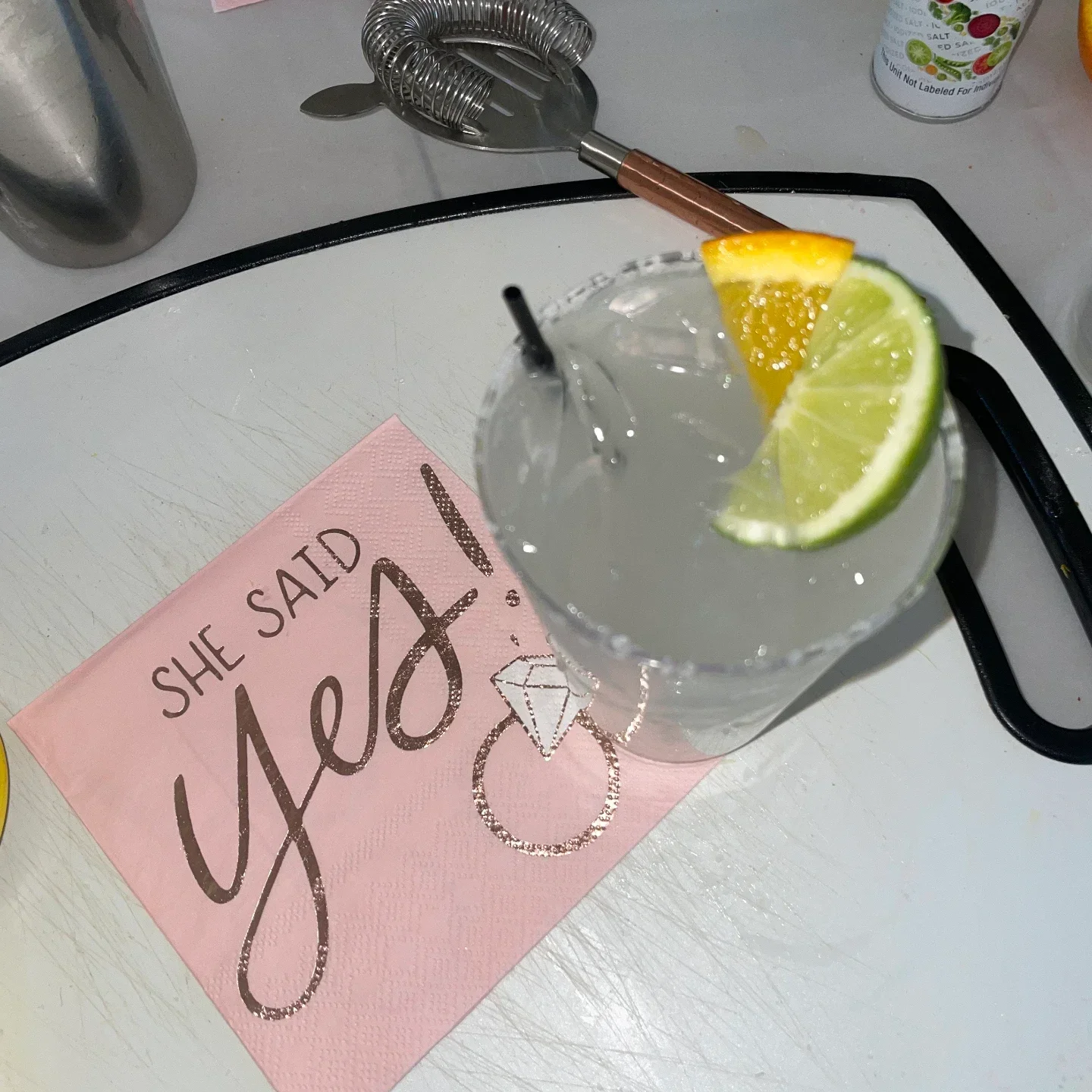 A rocks glass of clear cocktail with a salted rim, lime and orange wedges and a black straw, beside a pink napkin printed "SHE SAID yes!" with a diamond ring, on a white board with a strainer and shaker