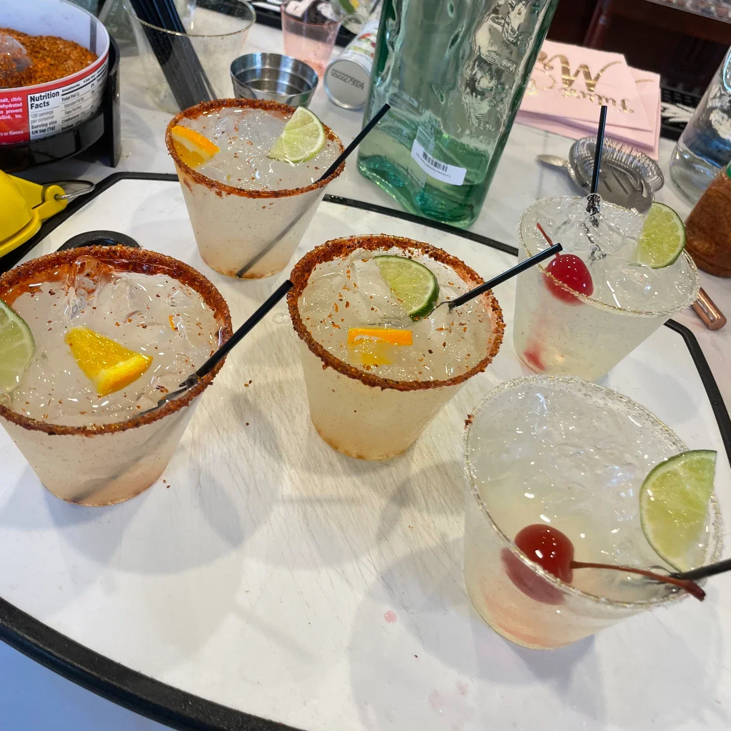 Five iced cocktails on a white board — three with chili rims, lime and orange, two with salt rims and bright red cherries — a bartender in a grey shirt behind the counter