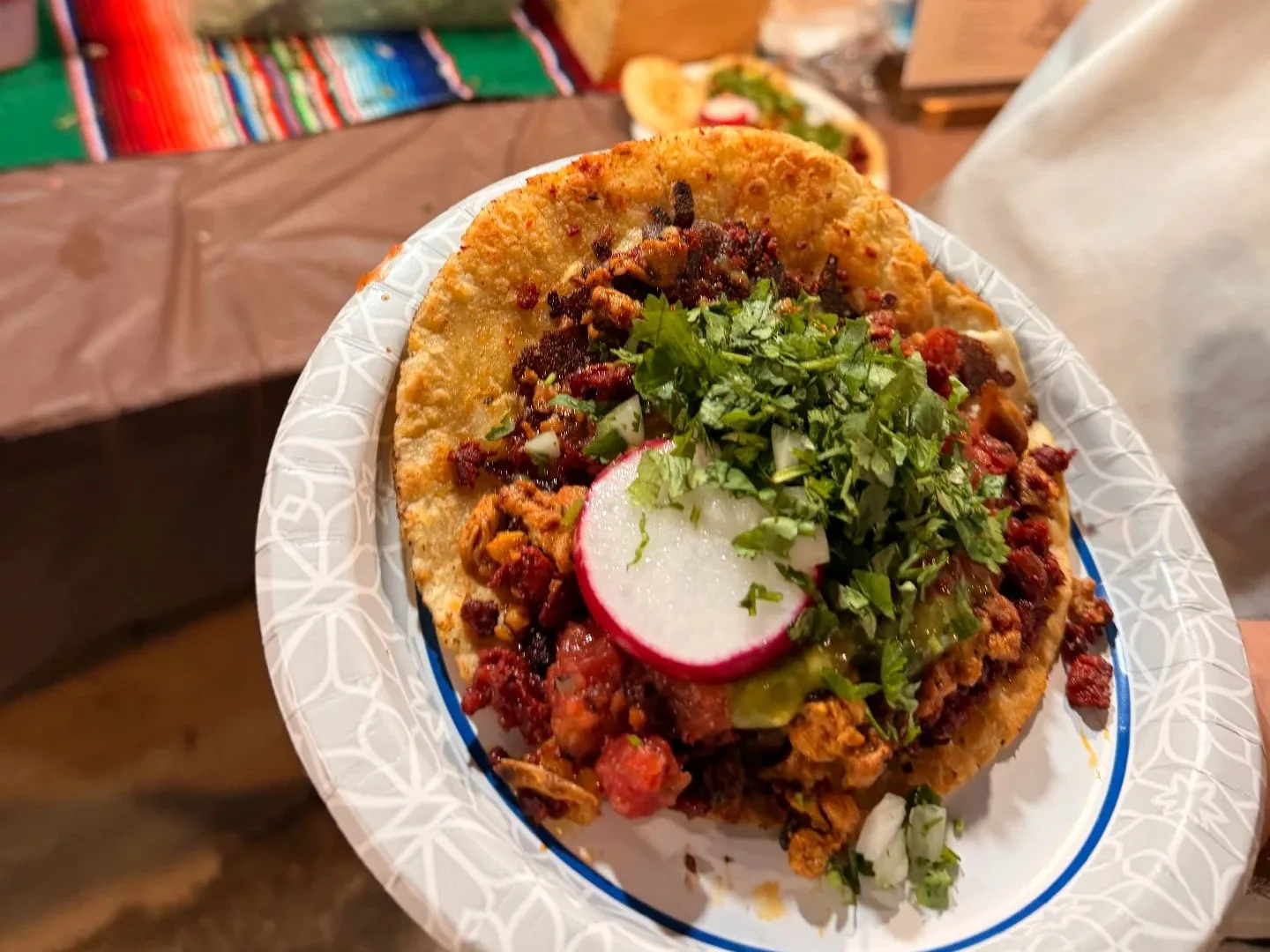 Taquiza: Crispy taco loaded with al pastor, chorizo, cilantro and a radish slice, held up beside a chili-rimmed cocktail with a paper umbrella