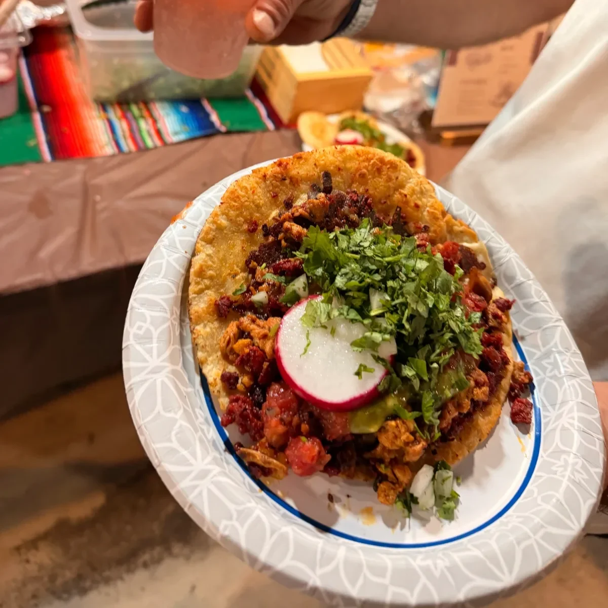 A guest holds a loaded taco with cilantro, onion and a radish slice on a paper plate, a chili-rimmed pink cocktail held just above