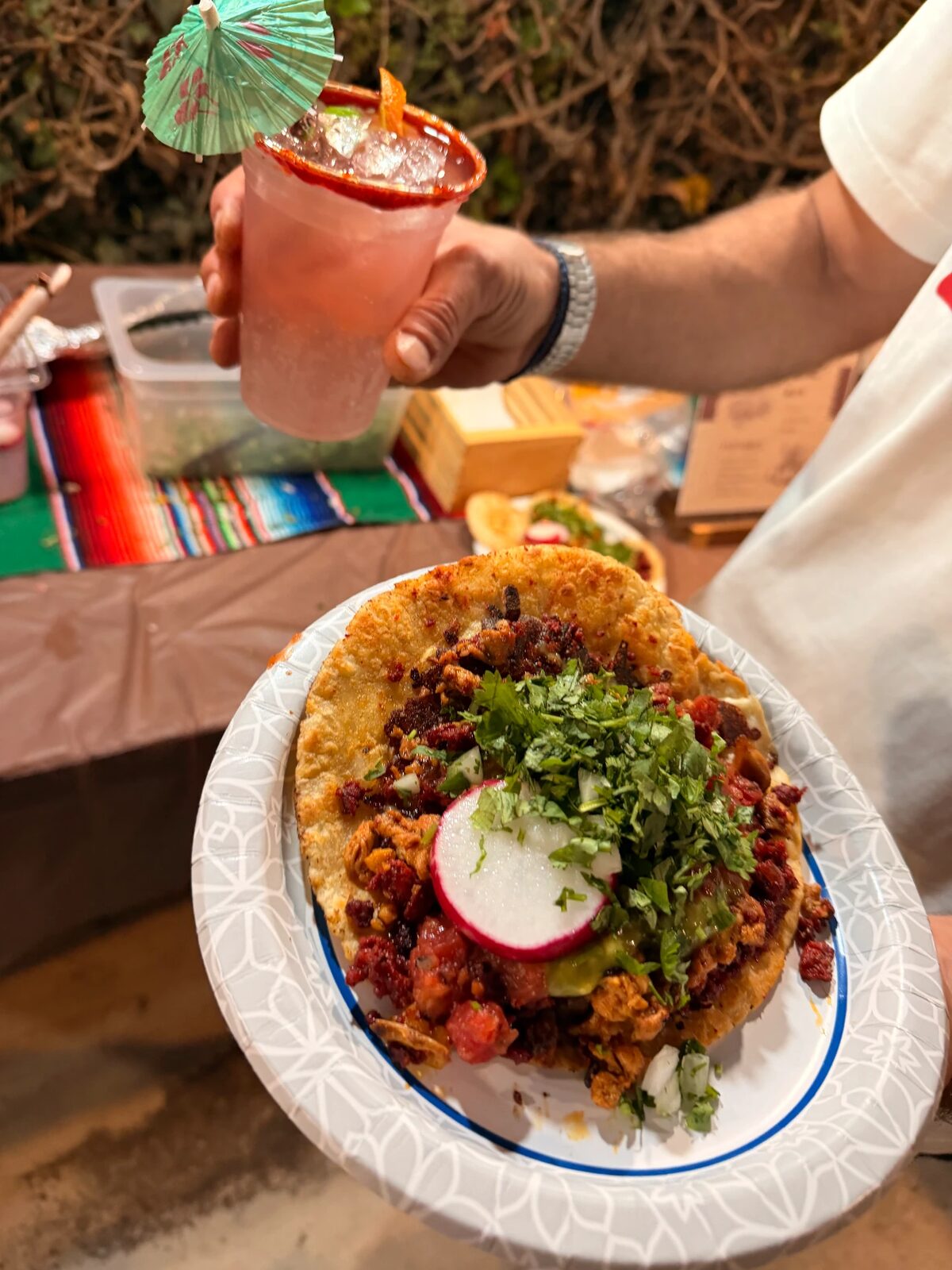 A guest holds a loaded taco topped with cilantro, onion and radish beside a chili-rimmed drink with a paper umbrella, over a sarape-covered serving table