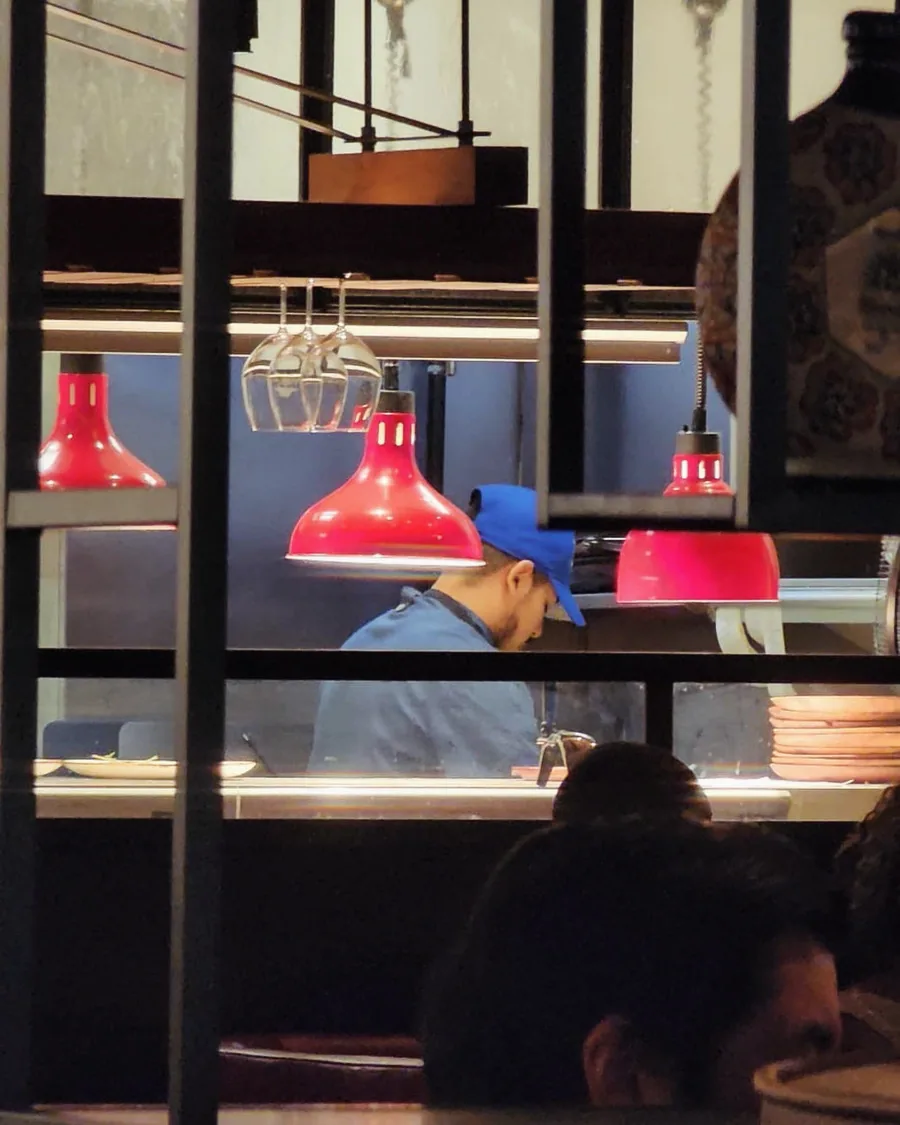A Pacheko cook in a blue cap working the kitchen pass under red heat lamps, with wine glasses hanging overhead