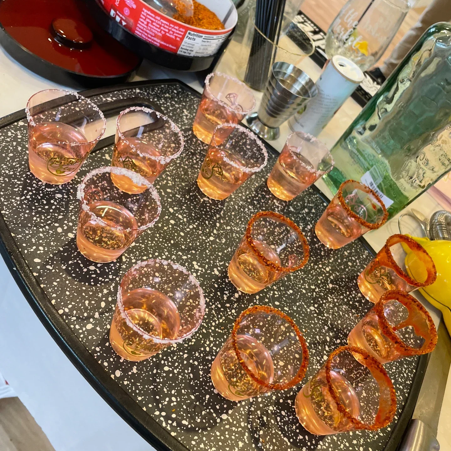 A row of shot glasses on a speckled board — half rimmed with salt, half with chili seasoning — filled with a pale amber pour, with a rimmer, jigger and tip jar behind